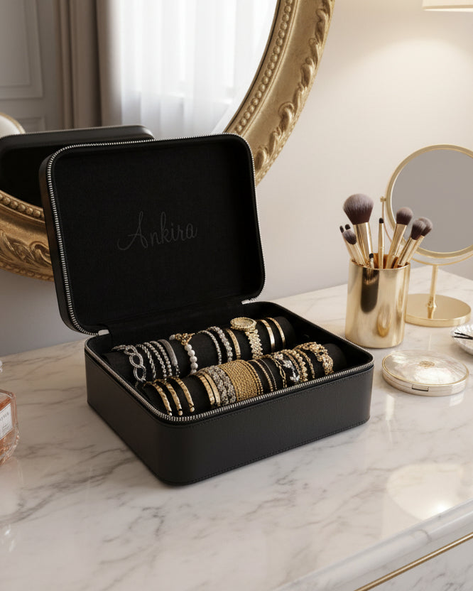 Black jewelry box with bracelets on a table next to makeup brushes and a mirror on a marble table